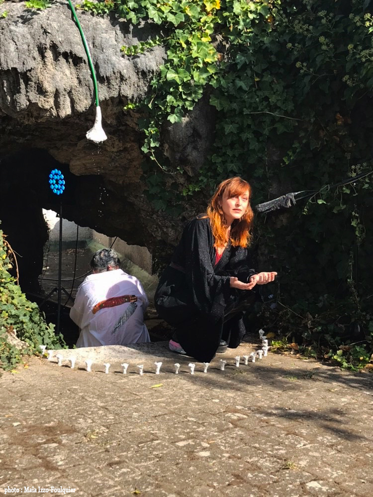On voi Sarah, l'artiste qui a créé le paysage sonore dans la performance, accroupie devant le micro. Clélia est de dos, sous le pommeau de couche qi coule, dans la grotte. Sur son imperméable on voit une peinture d'un couteau Laguiole. Un cercle de chanterelles les encercle.